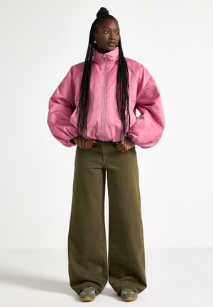 Kvinde med lange fletninger iført en skinnende lyserød lynlåstrøje, olivengrønne bukser med brede ben og grønne sneakers, stående med hænderne på hoften.