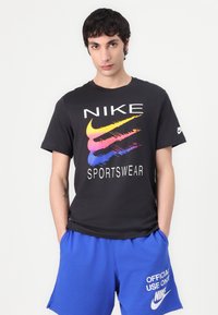 Tricou negru Nike cu un logo colorat și textul "SPORTSWEAR", asortat cu pantaloni scurți albaștri care afișează "FOLISIRE OFICIALĂ NUMAI" în alb.