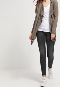 Lång, öppen kardigan i olivgrön, lager på lager över en figurnära vit t-shirt med V-ringning. Kombinerat med mörka skinny jeans och vita sneakers.