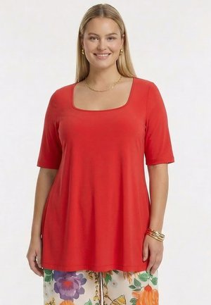 Smilende kvinde med langt blond hår iført en rød korteærmet top og farverige blomstrede bukser, accessoriseret med guldsmykker.