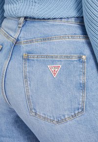 Lichtblauwe jeans hebben een driehoekig rood en wit "GUESS" logo patch op de achterzak, met zichtbare stiksels en textuur.