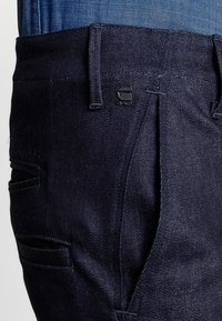 Dark denim pants featuring a straight cut, multiple pockets, and a sleek design with a small branded logo on the front waistband.