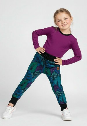 Młoda dziewczyna w fioletowej koszulce z długim rękawem i kolorowych spodniach z wzorem liści stoi z rękami na biodrach, uśmiechając się na prostym tle.