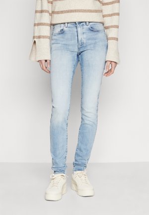 Jean skinny bleu clair porté avec des baskets beiges et un pull crème à rayures marron, debout sur un fond blanc.
