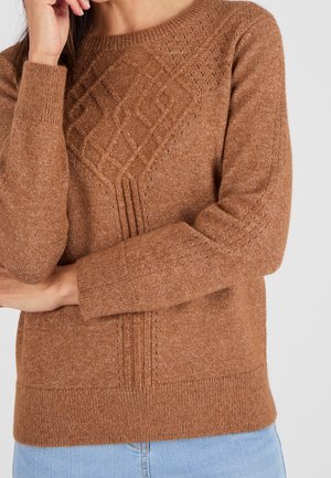 Pull marron tricoté avec motif losange texturé sur la poitrine, porté par une personne aux bras croisés et portant un jean bleu clair.