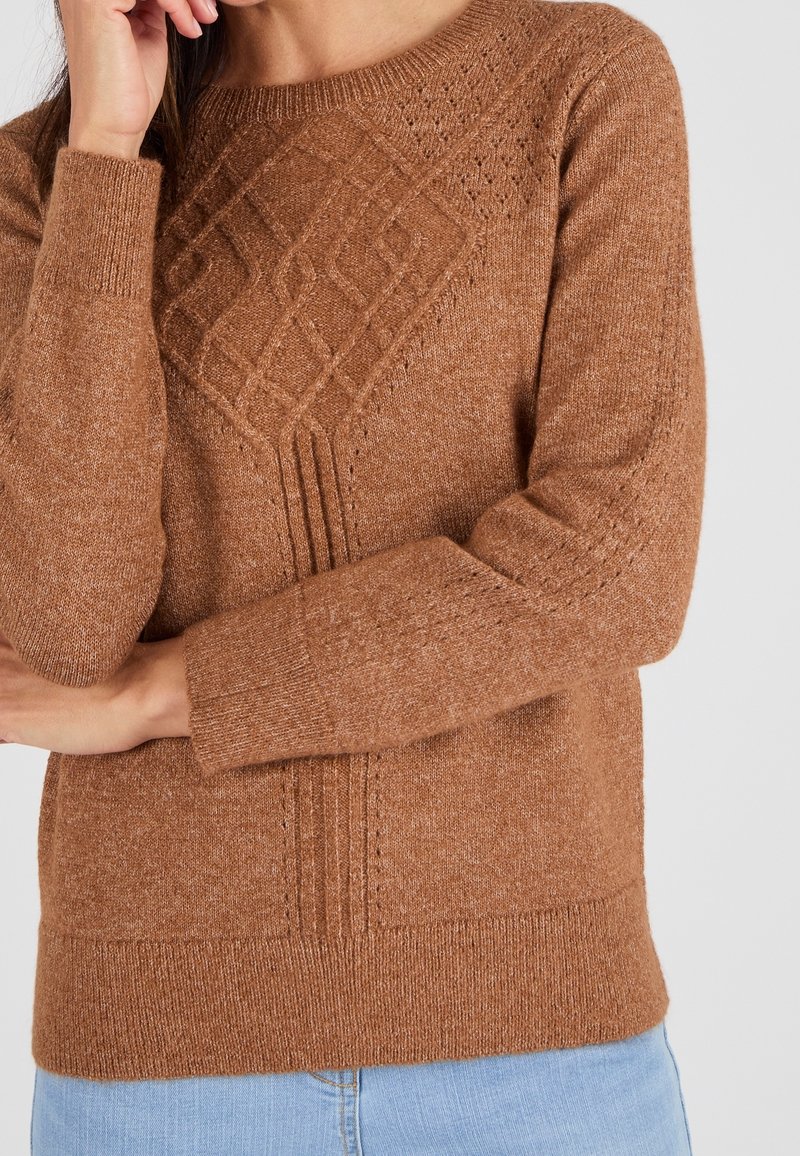 Pull marron tricoté avec motif losange texturé sur la poitrine, porté par une personne aux bras croisés et portant un jean bleu clair.