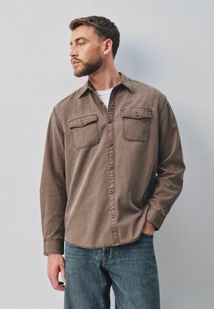 Mand med skæg iført en brun skjorte med knapper over en hvid t-shirt og blå jeans, står med den ene hånd i lommen og kigger mod venstre.
