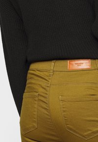 Spodnie w musztardowym kolorze, wykonane z mieszanki bawełny, mają tylne kieszenie i skórzaną metkę. Połączone z czarnym swetrem o teksturowanej fakturze.