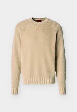 Beiger Strickpullover mit Struktur, langen Ärmeln, gerippten Bündchen und Saum sowie einem kleinen rechteckigen Logo-Patch auf der Brust.