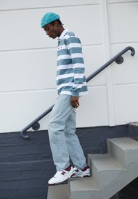 Chemise à rayures à manches longues en teal et blanc, jeans bleu clair et baskets New Balance avec des accents bordeaux, se tenant sur un escalier.