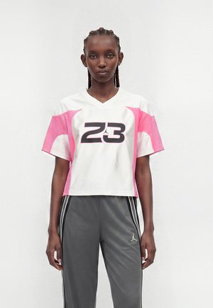 Femme aux cheveux tressés portant un maillot de sport blanc et rose avec le numéro 23 et un pantalon de survêtement gris avec des bandes blanches et un logo.