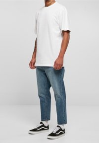 Hombre que lleva una camiseta blanca sencilla, jeans cortos azules, calcetines blancos y zapatillas negro y blancas, de pie contra un fondo simple.
