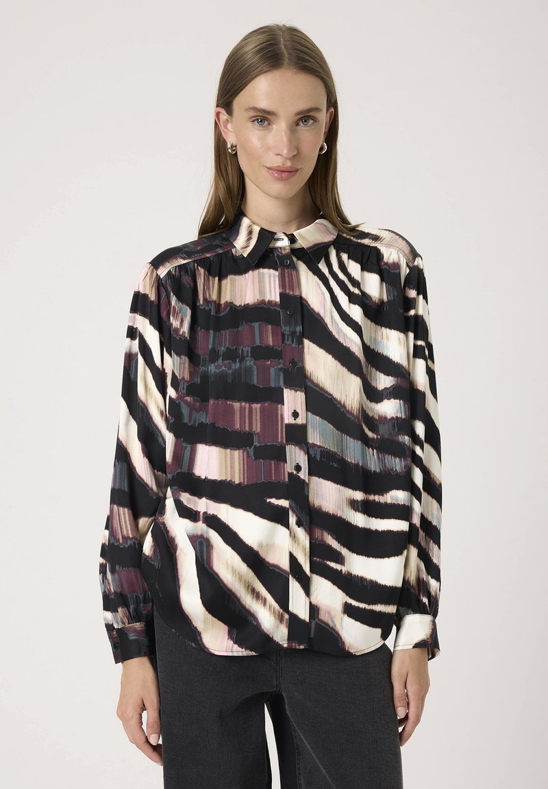 Kvinde iført en langærmet, knaplukket skjorte med et abstrakt zebraprint i sort, hvid, pink og blå nuancer, som er parret med mørke bukser.