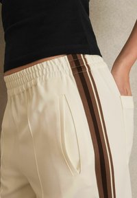 Krämfärgade joggers med bruna sidostänger, som bärs av en person i en svart topp, med fokus på midjan och fickorna.