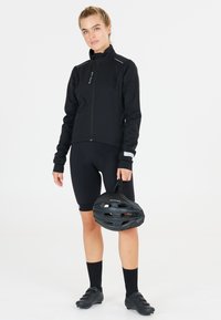 Zwarte sportjack met een hoge kraag, ritssluiting aan de voorkant en reflecterende details, gecombineerd met zwarte shorts, sokken en gripvaste fietsschoenen, terwijl hij een helm vasthoudt.