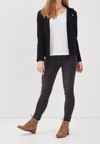 Donna in piedi che indossa una giacca nera, una camicia bianca, jeans skinny grigio scuro e stivali alla caviglia marroni con lacci su uno sfondo bianco.