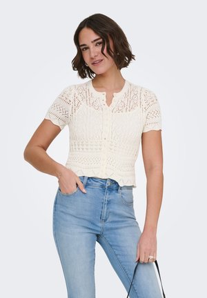 Joven con una blusa blanca de encaje con botones y mangas cortas y vaqueros de talle alto de color azul claro, sujetando una correa negra con una mano.