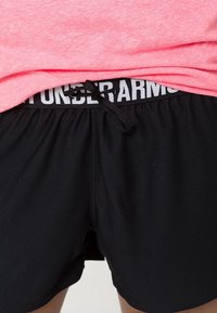 Svarta träningstights med dragsko, med ett texturerat midjeband där "UNDER ARMOUR" syns i kontrasterande vit text. Rosa skjorta ovanför.
