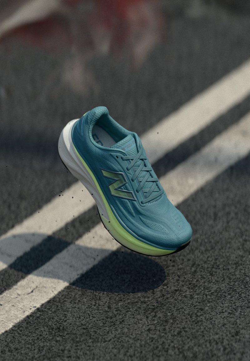 Μπλε παπούτσι τρεξίματος New Balance στον αέρα πάνω από έναν δρόμο με λευκές διαγραμμίσεις λωρίδων και σκορπισμένα μαύρα θραύσματα.