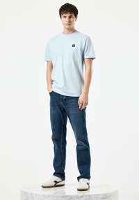 Lichtblauw T-shirt met korte mouwen en een zwart logo-embleem, gecombineerd met donkerblauwe jeans en witte sneakers met zwarte accenten, op een marmeren voetstuk.