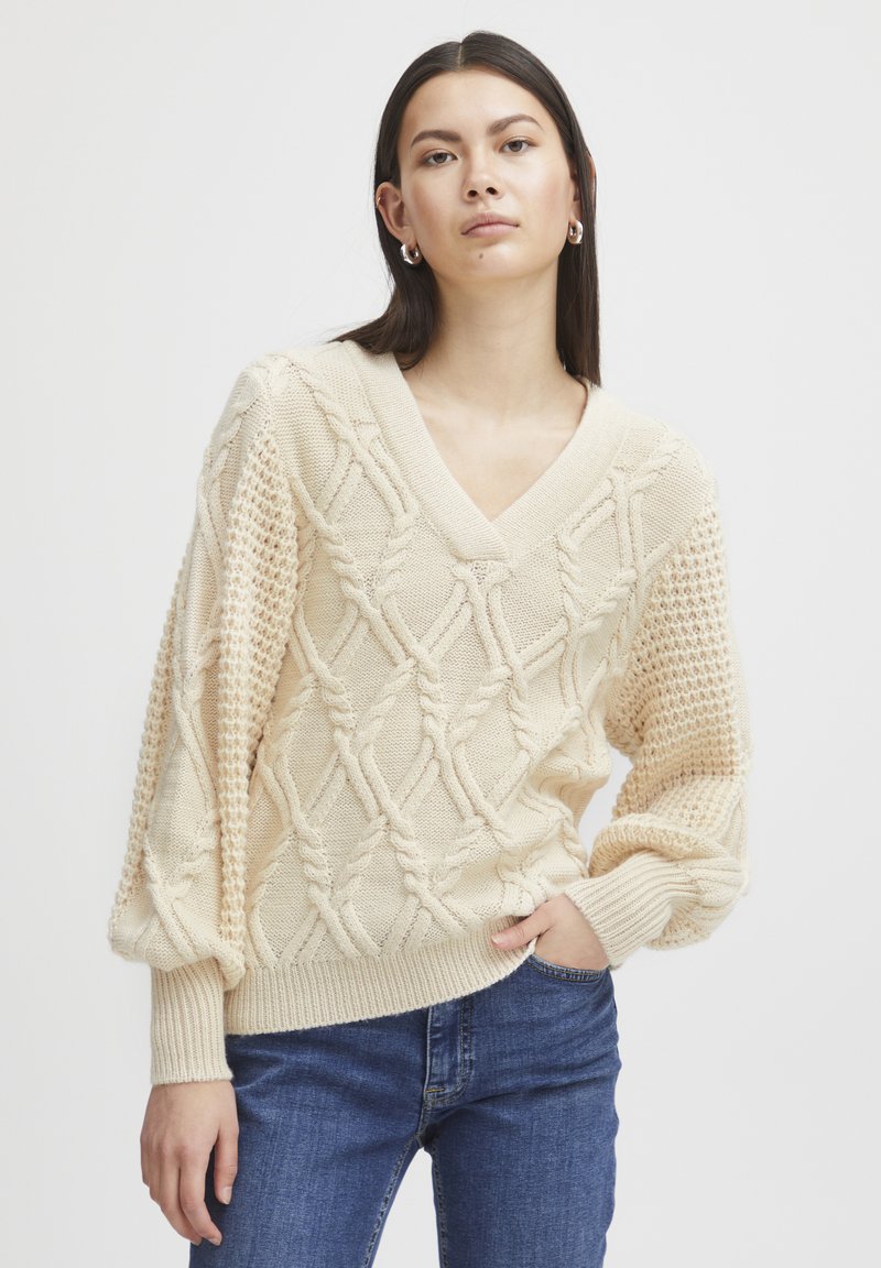 Pull en tricot crème avec un décolleté en V, des motifs texturés et des manches ballon. Poignets et ourlet côtelés. Associé à un jean bleu.