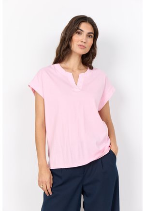 Femme aux longs cheveux bruns portant un haut rose pâle à manches courtes et un pantalon bleu marine, posant devant un fond blanc.