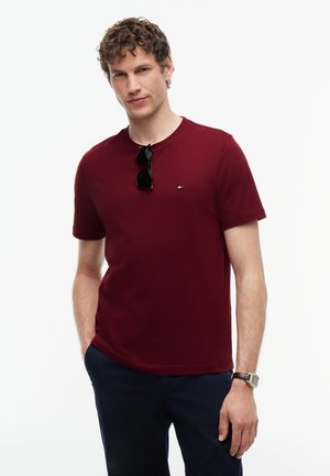 Uomo con capelli ricci che indossa una maglietta bordeaux, pantaloni scuri, occhiali da sole sul colletto e un orologio da polso, in piedi con una mano in tasca.