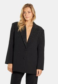 Blazer negro con escote en V profundo, adornado con tachuelas plateadas en los hombros, cierre de un solo botón y dos bolsillos delanteros. Tela texturada.