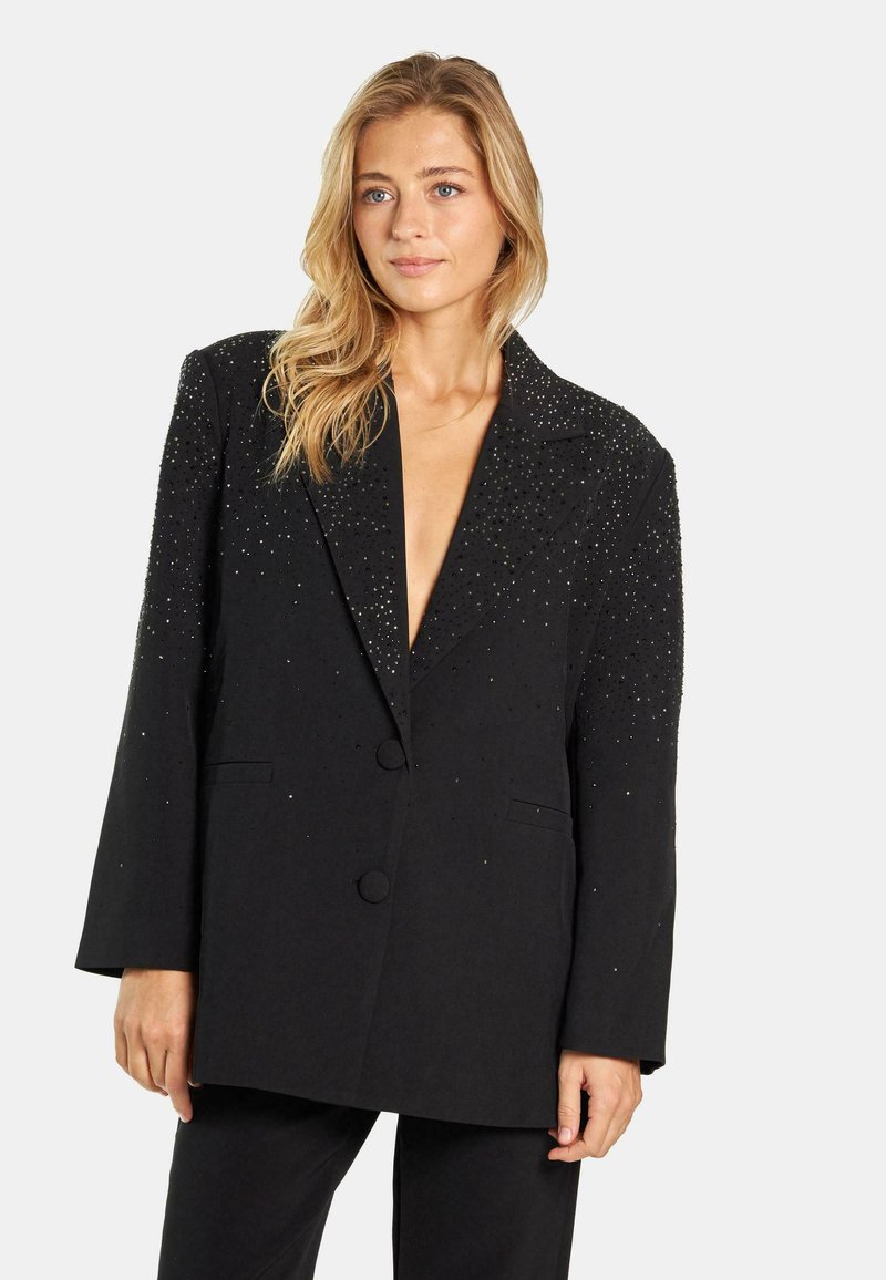 Blazer negro con escote en V profundo, adornado con tachuelas plateadas en los hombros, cierre de un solo botón y dos bolsillos delanteros. Tela texturada.