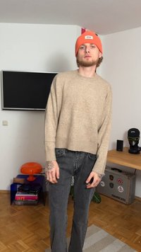 Suéter beige de punto con mangas anchas, combinado con jeans acampanados de color gris oscuro. Un gorro naranja añade un acento de color audaz. La habitación tiene suelo de madera.