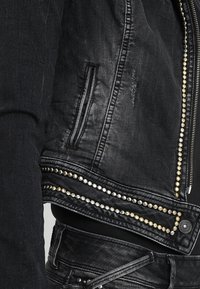 Detailní záběr na černou džínovou bundu s stříbrnými a zlatými hroty podél lemu a zipu, nošenou přes odpovídající černé džíny.