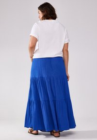 Gonna maxi a balze blu realizzata in tessuto leggero, abbinata a una t-shirt bianca con maniche arrotolate. Sandali visibili nella parte inferiore.