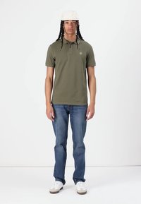 Olivgrön polo t-shirt med korta ärmar, klassisk krage och logotyp; ihop med blå jeans och vita sneakers. Enkelt bakgrund.