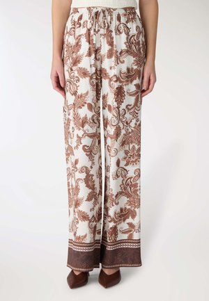Pantaloni bianchi a gamba larga con intricati motivi floreali e paisley marroni, vita con coulisse, indossati con scarpe marroni appuntite.