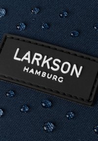 Sötétkék vászon anyag, kiemelkedő kék gyöngyökkel és egy fekete téglalap alakú címkével, melyen fehér betűkkel az áll: "LARKSON HAMBURG".