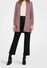 Langer hellrosa Veloursledermantel mit Stehkragen, kombiniert mit einem cremefarbenen Top und schwarzen Hosen, abgerundet mit schwarzen Stiefeletten mit Absatz.