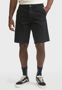 Black denim shorts with a straight cut, five pockets, and a front button closure. Paired with black sneakers and patterned socks.