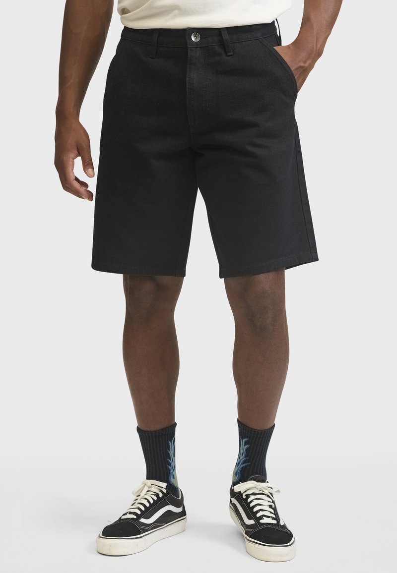 Black denim shorts with a straight cut, five pockets, and a front button closure. Paired with black sneakers and patterned socks.