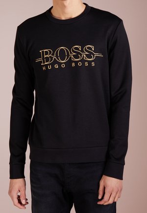 Musta pitkähihaisen collegepaita, jossa on kultainen "BOSS HUGO BOSS" -logo rinnassa, päällä henkilö, jolla on tummat housut.