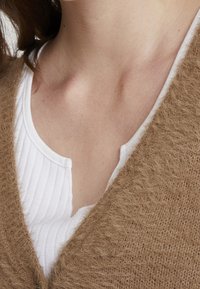 Brun stickad cardigan med en mjuk, fuzzy textur, lager på lager över en vit ribbad topp med knäppt halsringning.