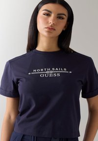 Jeune femme aux longs cheveux foncés portant un t-shirt bleu marine avec le logo « North Sails GUESS » sur un fond clair uni.