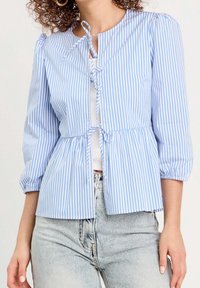 Femme portant un chemisier à rayures verticales bleues et blanches avec des manches trois-quarts et deux nœuds à l'avant, par-dessus un haut blanc et un jean en denim clair.