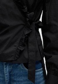 Schwarze Bluse mit Rüschensaum, an der Taille gebunden, kombiniert mit blauen Denim-Jeans, die die Detailansicht der Ärmelbündchen und der Taschen zeigen.