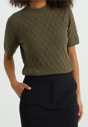 Vrouw met een olijfgroen textuurrijk kortmouwig truitje en zwarte hoge taille broek, één hand in de zak, met zichtbaar krullend haar.