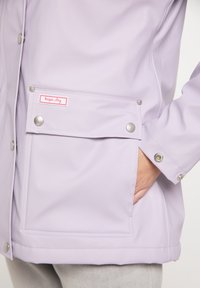 Giacca leggera color lavanda realizzata in materiale waterproof liscio. Presenta una tasca frontale, bottoni a pressione argentati e un'etichetta rosa "mantiene asciutto".