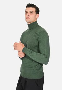 Maglione verde a collo alto realizzato in tessuto morbido e testurizzato; design aderente con maniche lunghe e polsini a coste; abbinato a pantaloni neri.