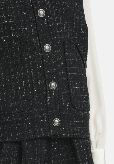 Gilet en tweed noir avec boutons métalliques, surface texturée et accents brillants ; comprend deux poches avant et une blouse blanche en dessous.