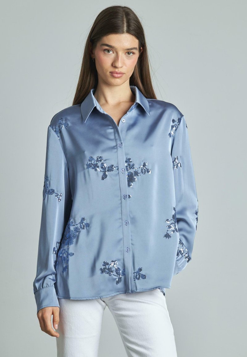 Camicia azzurra in satin con bottoni, decorata con applicazioni di paillettes floreali, maniche lunghe e colletto classico. Tessuto morbido e vestibilità rilassata.