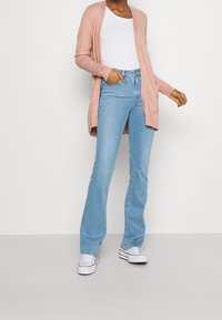 Mujer vestida con jeans de corte bootcut de color azul claro, zapatillas blancas, blusa blanca y un cárdigan rosa abierto, de pie con una mano en el bolsillo.