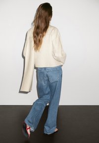 Cardigan en mélange de laine beige, coupe ample et manches longues, associé à un jean taille haute bleu clair, porté avec des sandales noires à semelles rouges.